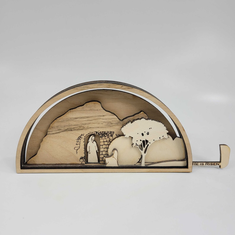 May include: A wooden, half-circle sculpture depicting a scene from the Bible. The sculpture is made of light brown wood and features a carved image of a woman standing in front of a stone tomb. The tomb has a stone door with the word 'Christ' carved above it. A tree is to the right of the tomb. The sculpture is mounted on a wooden base with the words 'He is Risen' carved into the base.