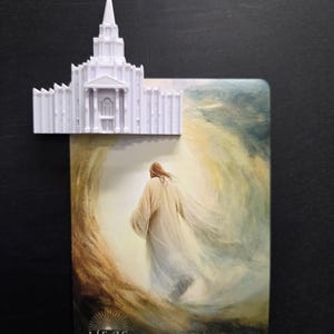 May include: A white 3D-printed temple model attached to a rectangular card featuring an illustration of a figure in a robe walking toward a bright light. The text HE IS RISEN MATTHEW 28:6-7 is printed in white at the bottom of the card.