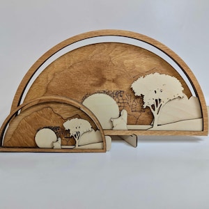 May include: A wooden wall decor with a cutout design of a landscape with trees and a stone archway. The decor is made of two pieces, a larger piece with a semi-circle shape and a smaller piece with a semi-circle shape.