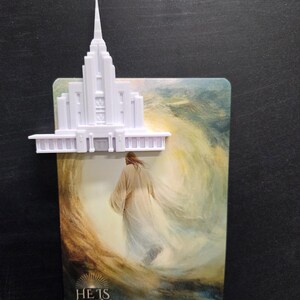 May include: A white 3D-printed temple structure mounted on a rectangular card featuring an illustration of a figure in a robe walking toward a bright light, with the text HE IS RISEN MATTHEW 28:6-7 printed at the bottom.