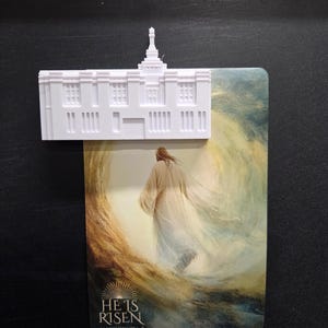 May include: A white 3D-printed model of a temple building positioned atop a rectangular card. The card features an illustration of a figure in a robe walking toward a bright light, with the text HE IS RISEN and MATTHEW 28:6-7 printed at the bottom.