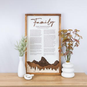 May include: A framed wooden sign with the word "Family" in a cursive font. The sign includes text and a mountain and tree line design. Two white vases with flowers and a white ceramic knot are on a wooden surface.