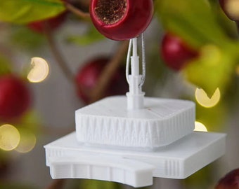 Op de afbeelding: Witte miniatuurgebouwornament met een decoratief bovenstuk, hangend aan een tak met rode bessen.