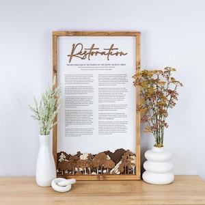 May include: Wooden framed wall art with the word "Restoration" in a script font. The text below reads "The Restoration of the Fullness of the Gospel of Jesus Christ." A forest scene is at the bottom. Two white vases with flowers are on either side.