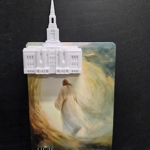 May include: A white plastic temple-shaped ornament attached to a rectangular card featuring an illustration of a figure in a robe walking towards a bright light. The text at the bottom reads HE IS RISEN MATTHEW 28:6-7 in gold lettering.