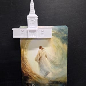 May include: A white plastic church building model attached to a rectangular card featuring an illustration of a figure in a robe walking toward a bright light, with the text HE IS RISEN MATTHEW 28:6-7 printed at the bottom.