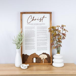 May include: Wooden framed wall art with the word "Christ" in a script font. The artwork features text and a landscape scene with a tomb and tree. The frame is brown, and the background is white. Decorative vases with flowers are on the table.