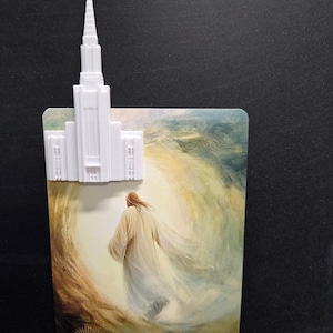May include: A white 3D-printed temple spire topper attached to a rectangular card featuring an illustration of a figure in a robe walking toward a bright light, with the text HE IS RISEN MATTHEW 28:6-7 printed at the bottom.