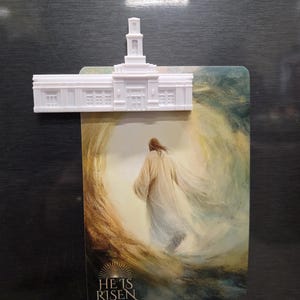 May include: A white plastic magnet shaped like a temple building holding a rectangular card. The card features an illustration of a figure walking toward a bright light with the text HE IS RISEN MATTHEW 28:6-7 printed at the bottom.
