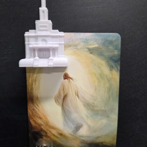 May include: A white 3D-printed temple topper attached to a rectangular card featuring an illustration of a figure in a robe walking toward a bright light, with the text "HE IS RISEN" and "MATTHEW 28:6-7" printed at the bottom of the card.