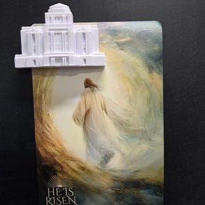 May include: A white 3D-printed temple model attached to a rectangular card featuring an illustration of a figure in a white robe walking toward a bright light. The text HE IS RISEN MATTHEW 28:6-7 is printed in white at the bottom of the card.