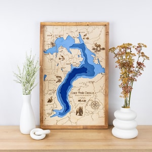 May include: A wooden map of Lake Pend Oreille, Idaho, with blue water and brown land features. The map is framed in a natural wood frame and includes the lake's name and coordinates.