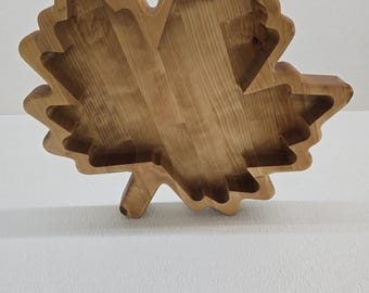 May include: A wooden maple leaf-shaped serving tray. The tray is made of light brown wood and has a carved design with multiple levels. The leaf's edges are detailed, and the tray is set against a white background. A decorative piece for serving snacks or displaying items.
