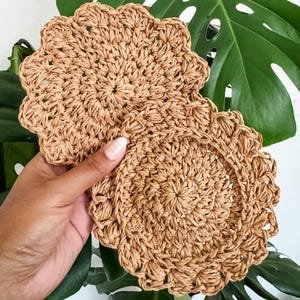 May include: Two round, woven coasters in a light brown color. The coasters have a scalloped edge and a textured, radial pattern. The background features a large, green tropical leaf.