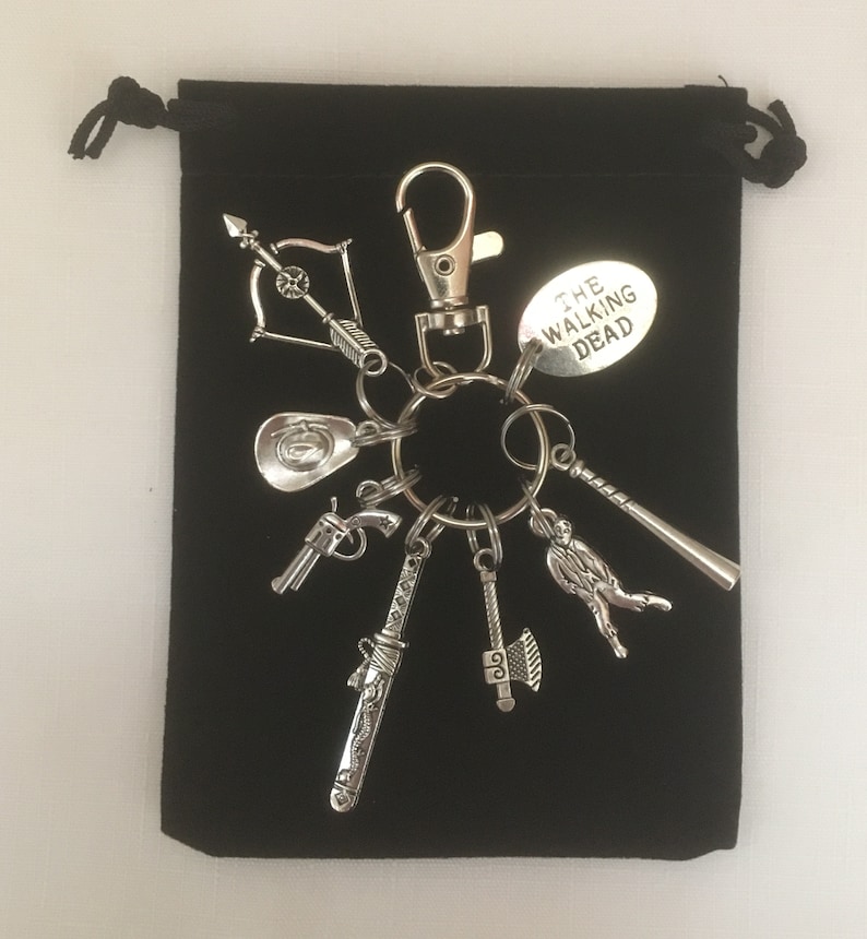 May include: Silver-coloured keychain with various charms, such as a bow and arrow, cowboy hat, pistol, sword, axe, baseball bat, and a charm reading "THE WALKING DEAD." Presented on a black drawstring bag.