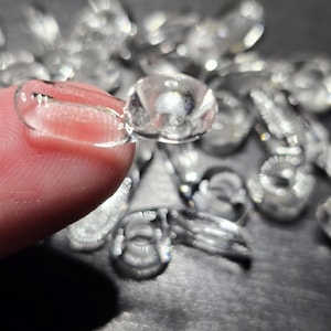 May include: Close-up of a clear, round contact lens held between fingers. The lens is transparent with a slight curve, reflecting light. Other lenses are scattered in the background, suggesting a collection or supply of contact lenses.