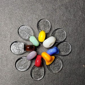 Op de afbeelding: Een set van tien glazen kralen in verschillende kleuren, waaronder rood, oranje, geel, groen, blauw, paars en wit. De kralen zijn in een cirkel op een grijs oppervlak gerangschikt.