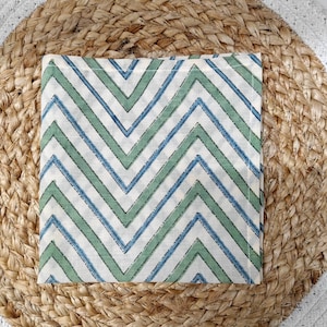 May include: A square, off-white cloth napkin with a repeating chevron pattern in blue and green. The napkin is placed on a round, woven jute placemat with a white border. The design is simple and modern.