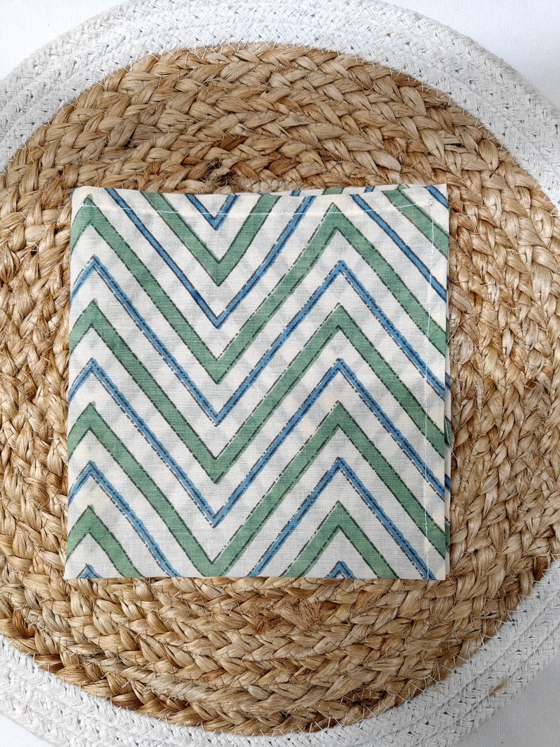 May include: A square, off-white cloth napkin with a repeating chevron pattern in blue and green. The napkin is placed on a round, woven jute placemat with a white border. The design is simple and modern.