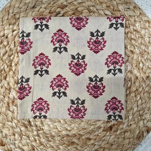 May include: A square, cream-colored fabric with a repeating floral pattern in shades of pink and dark gray. The flowers have multiple petals and are arranged in a symmetrical design. The fabric is placed on a woven, natural-toned surface.
