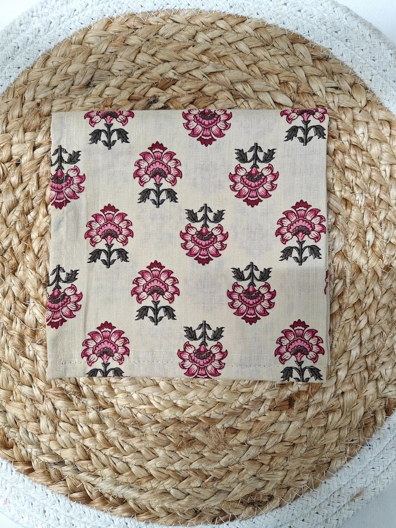 May include: A square, cream-colored fabric with a repeating floral pattern in shades of pink and dark gray. The flowers have multiple petals and are arranged in a symmetrical design. The fabric is placed on a woven, natural-toned surface.