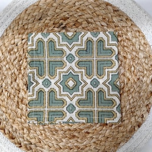 May include: A square, patterned fabric in shades of green and gold, placed on a round woven jute mat with a white border. The fabric features a geometric design with star-like shapes.