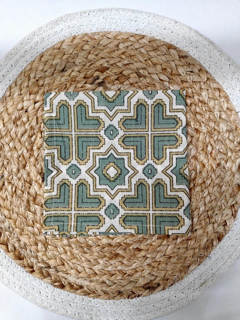 May include: A square, patterned fabric in shades of green and gold, placed on a round woven jute mat with a white border. The fabric features a geometric design with star-like shapes.