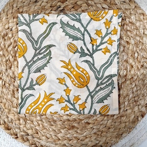 May include: Square white cloth napkin with a floral pattern in yellow and green. The napkin features a repeating design of stylized flowers and leaves. The napkin is placed on a round woven placemat.