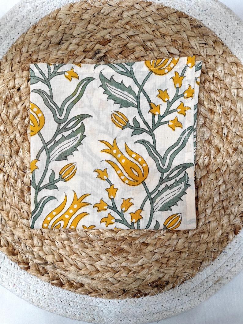 May include: Square white cloth napkin with a floral pattern in yellow and green. The napkin features a repeating design of stylized flowers and leaves. The napkin is placed on a round woven placemat.