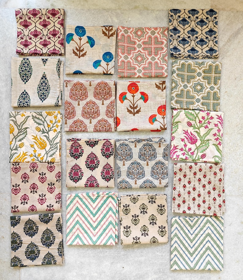 May include: A collection of folded square fabric napkins with various floral and geometric patterns. The napkins feature designs in shades of blue, red, pink, green, and yellow on a neutral background. The patterns include stylized flowers, trees, and geometric shapes.