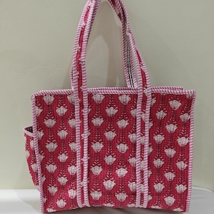 May include: A quilted tote bag with a floral pattern in shades of red and white. The bag features a rectangular shape with a side pocket and sturdy handles. The edges and handles are trimmed with a pink and white striped fabric.