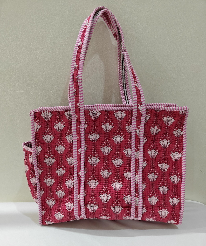 May include: A quilted tote bag with a floral pattern in shades of red and white. The bag features a rectangular shape with a side pocket and sturdy handles. The edges and handles are trimmed with a pink and white striped fabric.