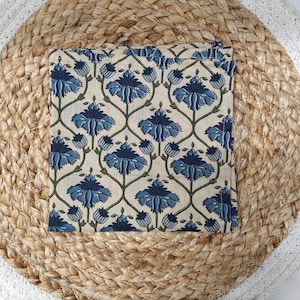 May include: A folded square textile with a repeating floral pattern in shades of blue and green on an off-white background. The textile is centered on a round, woven placemat with a natural fiber appearance and a white border.