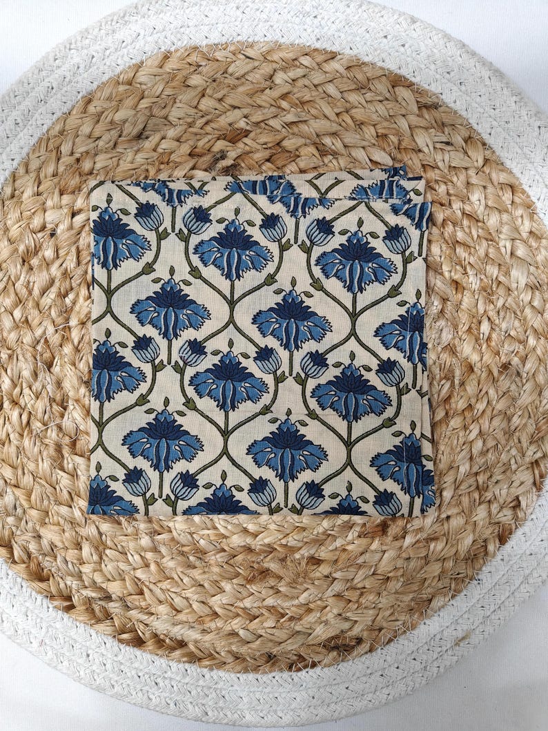 May include: A folded square textile with a repeating floral pattern in shades of blue and green on an off-white background. The textile is centered on a round, woven placemat with a natural fiber appearance and a white border.