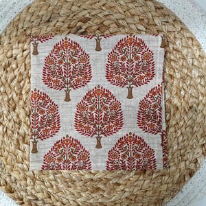 May include: A square, off-white fabric with a repeating tree pattern in shades of red, orange, and brown. The trees have detailed leaf and flower designs. The fabric is placed on a woven, circular mat.