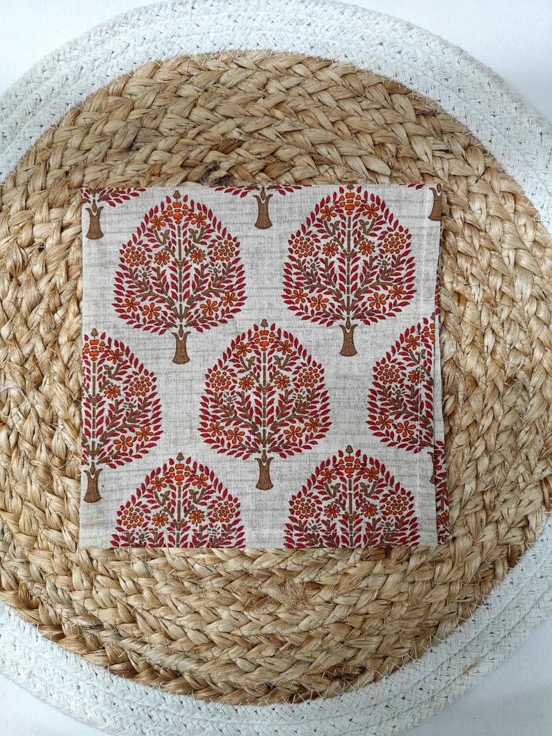 May include: A square, off-white fabric with a repeating tree pattern in shades of red, orange, and brown. The trees have detailed leaf and flower designs. The fabric is placed on a woven, circular mat.