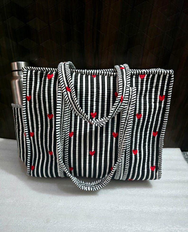 May include: Black and white striped tote bag with red heart accents. The bag has a water bottle pocket and matching striped handles. The tote is rectangular and appears to be made of quilted fabric.