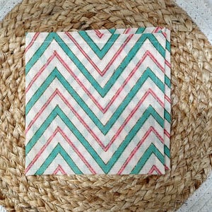 May include: Square cloth napkin with a chevron pattern in teal and pink on a white background. The napkin is placed on a round woven placemat with a white border.