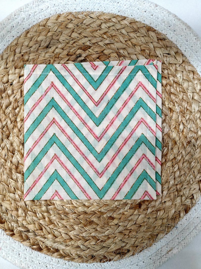 May include: Square cloth napkin with a chevron pattern in teal and pink on a white background. The napkin is placed on a round woven placemat with a white border.
