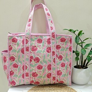 May include: A pink floral tote bag with a matching side pocket and handles. The bag features a repeating pattern of red flowers on a light pink background. The handles and edges are trimmed with a striped pink fabric.