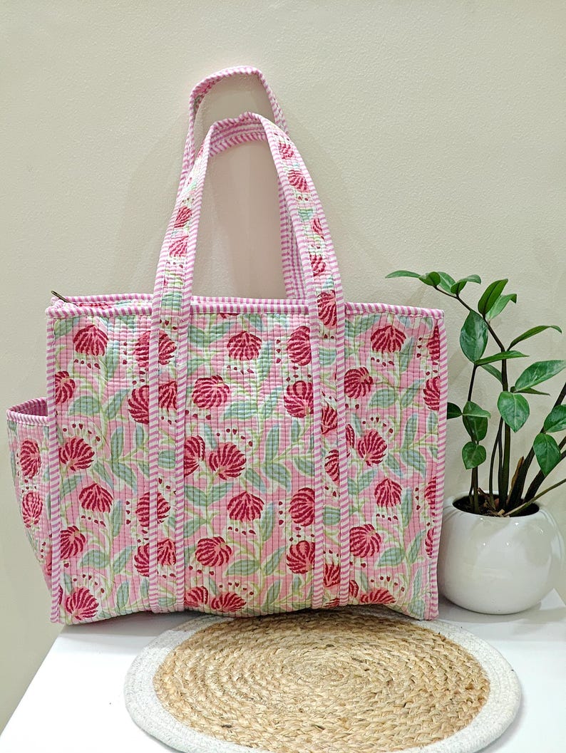May include: A pink floral tote bag with a matching side pocket and handles. The bag features a repeating pattern of red flowers on a light pink background. The handles and edges are trimmed with a striped pink fabric.