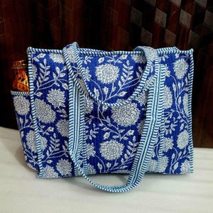 May include: A blue tote bag with a white floral pattern. The bag has a rectangular shape with a pocket on the side. The handles and trim are a lighter shade of blue. The bag appears to be made of quilted fabric.
