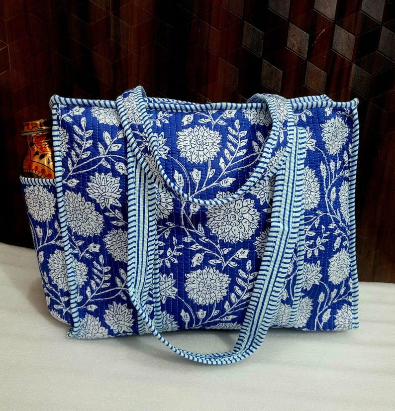 May include: A blue tote bag with a white floral pattern. The bag has a rectangular shape with a pocket on the side. The handles and trim are a lighter shade of blue. The bag appears to be made of quilted fabric.