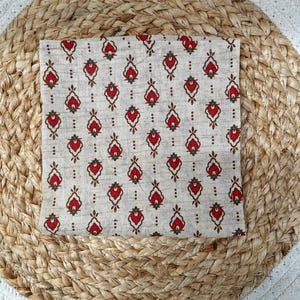 May include: A folded square of fabric with a repeating pattern of red and brown floral-like shapes on a beige background. The fabric is placed on a round woven mat with a white border.