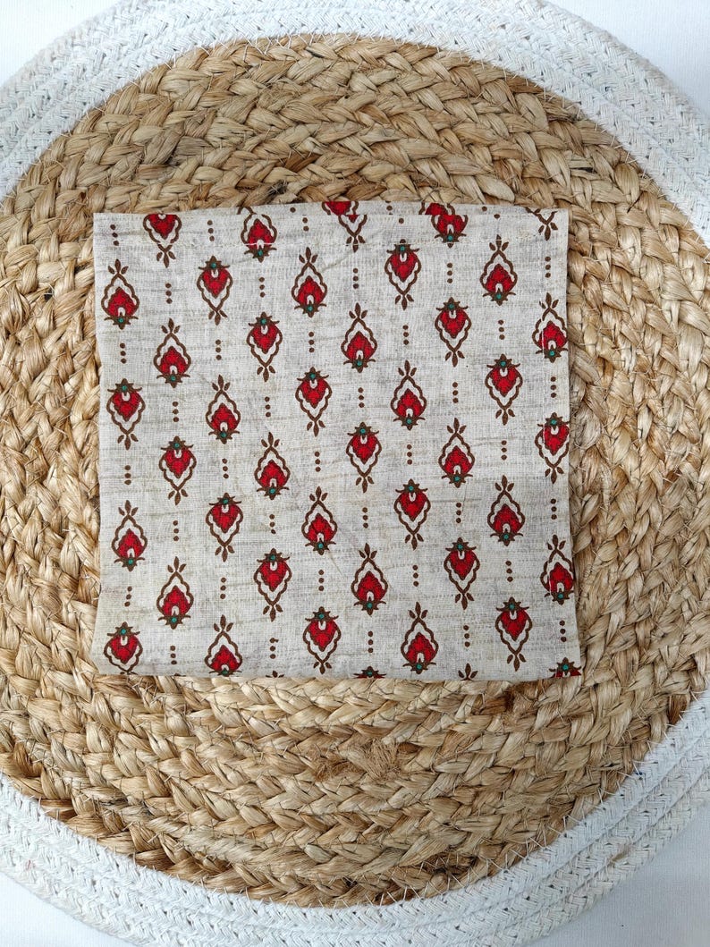 May include: A folded square of fabric with a repeating pattern of red and brown floral-like shapes on a beige background. The fabric is placed on a round woven mat with a white border.