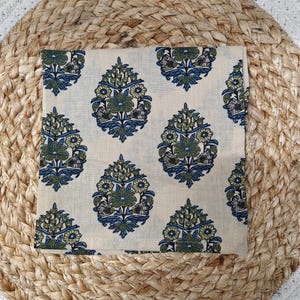 May include: A square, cream-colored cloth napkin with a repeating floral pattern in blue and green. The napkin is placed on a woven, beige placemat.