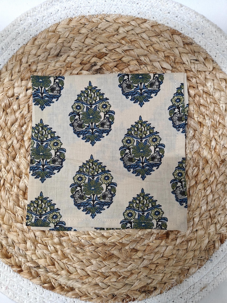 May include: A square, cream-colored cloth napkin with a repeating floral pattern in blue and green. The napkin is placed on a woven, beige placemat.