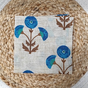 May include: A square, folded cloth napkin with a floral pattern. The design features blue flowers with teal accents and brown stems on a light beige background. The napkin is resting on a woven jute placemat.