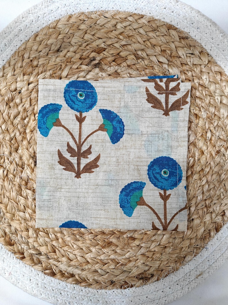 May include: A square, folded cloth napkin with a floral pattern. The design features blue flowers with teal accents and brown stems on a light beige background. The napkin is resting on a woven jute placemat.