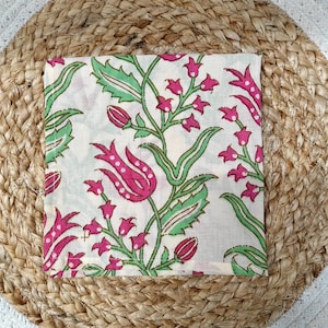 May include: Square white fabric with a floral pattern featuring pink flowers and green leaves. The fabric is placed on a round woven jute placemat with a white border. The floral design includes stylized tulips and smaller flower clusters.